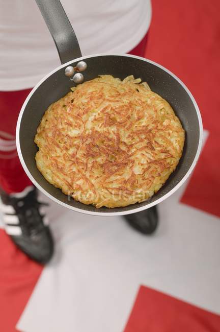Vista de primer plano del futbolista en la bandera suiza sosteniendo sartén con tortita de papa frita - foto de stock