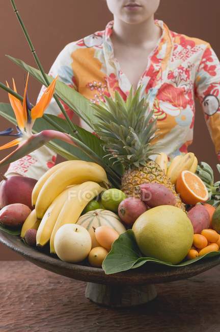 Cropped view of woman holding bowl of exotic fruits on brown background — Stock Photo