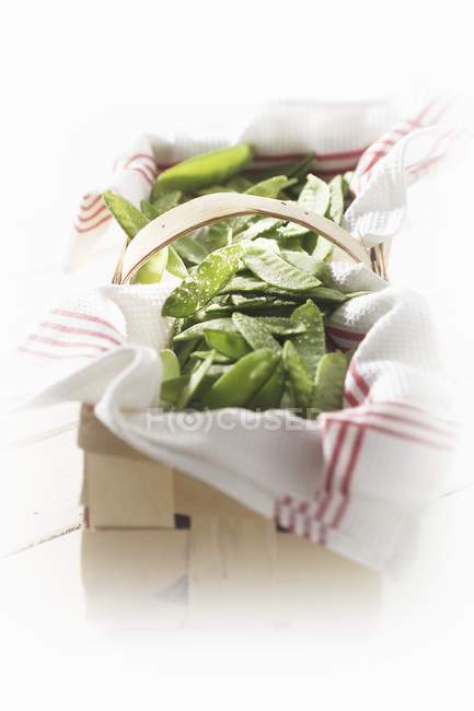 Fresh mangetout on tea towel in woodchip basket over white surface — Stock Photo