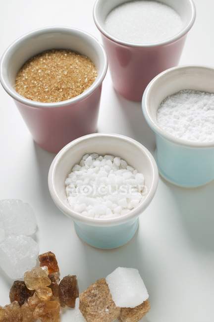 Primer plano vista elevada de varios tipos de azúcar en tazas y en la superficie blanca - foto de stock