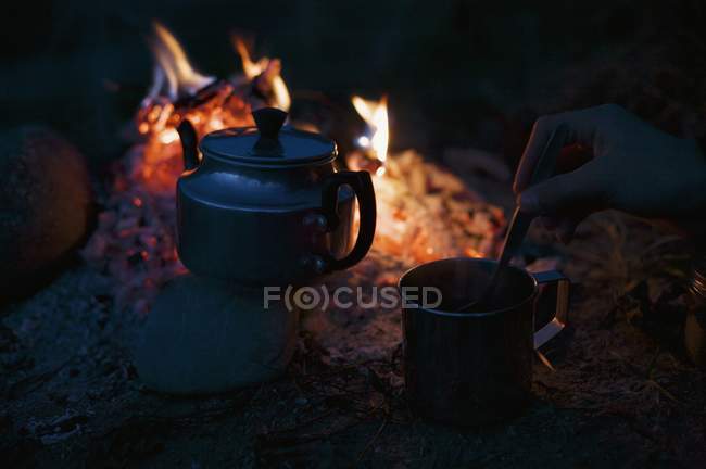 Mão mexendo chá na xícara por bule perto de fogo acampamento — Fotografia de Stock