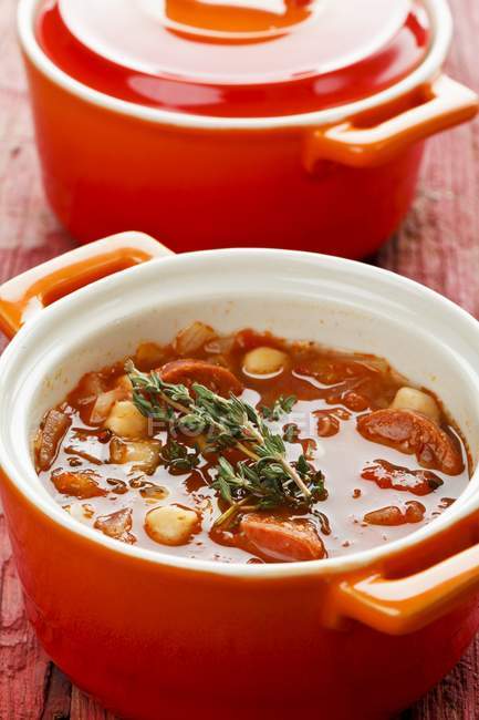 Vegetable stew with chickpeas and sausage in red pots over wooden surface — Stock Photo