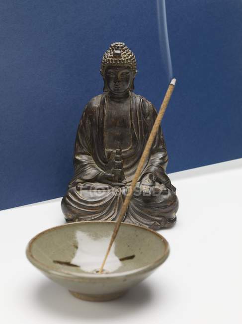 Elevated view of lit smoking incense stick in ceramic dish in front of Buddha figure — Stock Photo