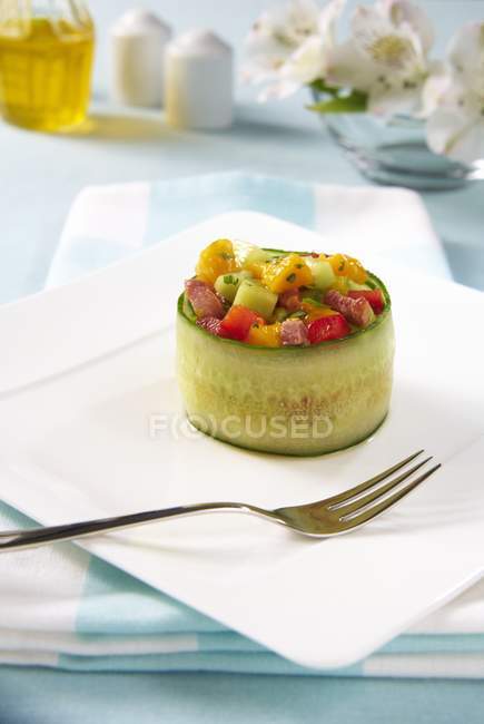 Un rollo de pepino relleno con ensalada de mango, pimienta, pepino y tocino en un plato blanco con tenedor - foto de stock