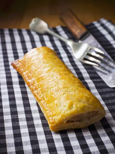 Closeup view of one sausage roll with fork and knife on checkered towel — Stock Photo