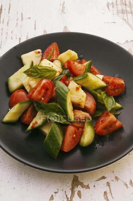 Tomato and cucumber salad with pineapple and basil on black plate  over wooden surface — Stock Photo