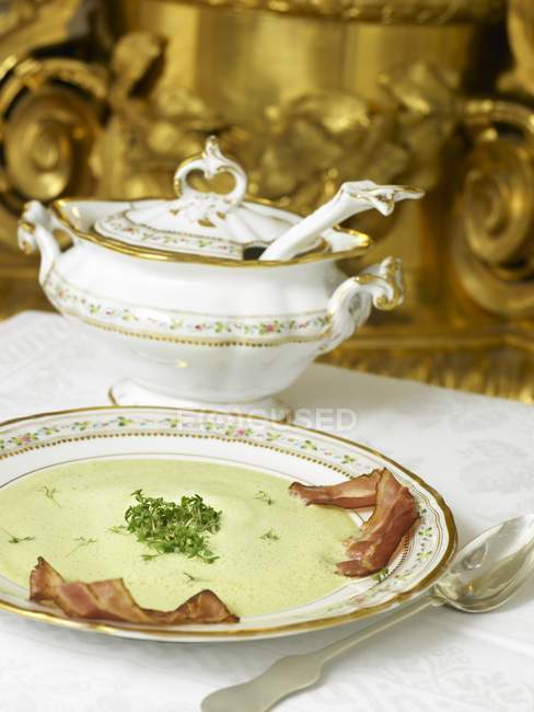 Cucumber soup with bacon and cress on white plate over towel with spoon — Stock Photo