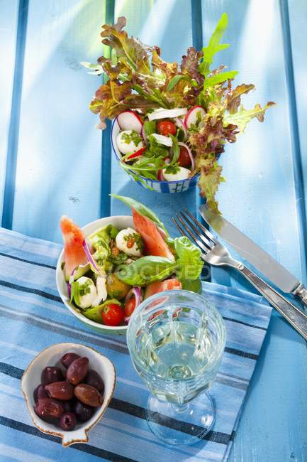 Two summer salads, olives and a glass of white wine over blue surface — Stock Photo