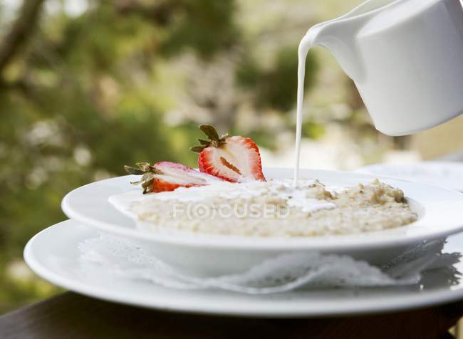 Vista close-up de leite derramando de um jarro para uma tigela de aveia com morangos — Fotografia de Stock