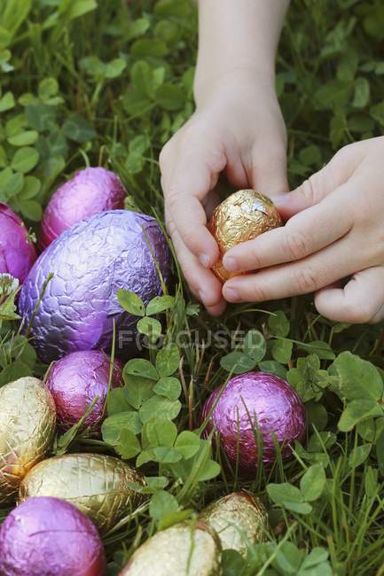 Primer plano vista recortada de las manos del niño sosteniendo huevo de chocolate envuelto en papel de aluminio en la hierba - foto de stock