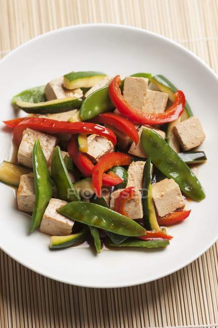 Remover-frito cubos de tofu y verduras sarna tout, calabacín y pimientos rojos con salsa de soja en plato blanco - foto de stock