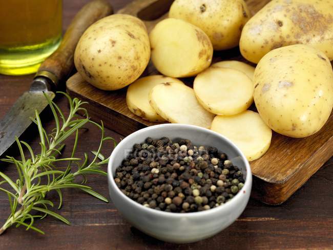 Potatoes, peppercorns and rosemary on chopping board and in bowl over wooden surface — Stock Photo