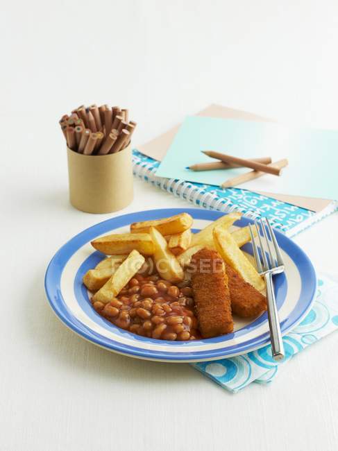 Рыбные пальцы с запеченными бобами и чипсами на голубой тарелке над белой поверхностью — стоковое фото