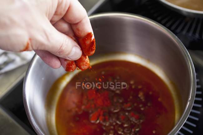Vista recortada de una mano poniendo pimentón en polvo a una olla de salsa - foto de stock