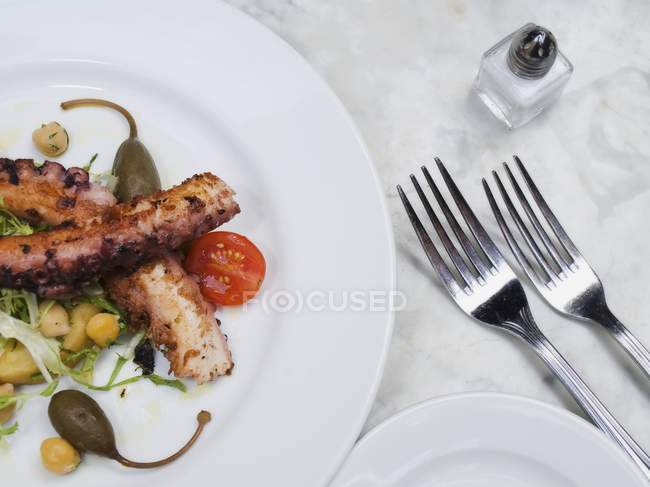 Жареный осьминог с помидорами черри, гигантскими каперсами и горохом на белой тарелке за столом с вилками — стоковое фото