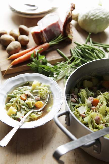 Sopa de verduras con ingredientes en plato con cuchara en cacerola sobre mesa de madera - foto de stock