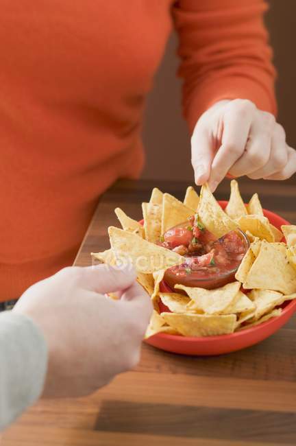 Hands dipping nachos in tomato salsa — Stock Photo