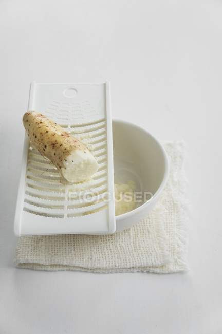 Rafano su una grattugia e una ciotola di rafano grattugiato su superficie bianca — Foto stock