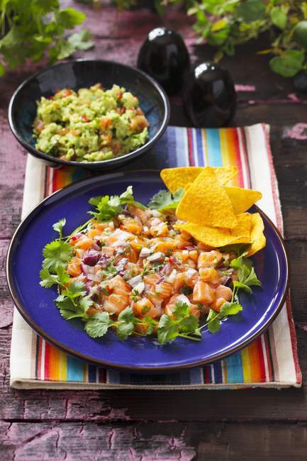 Ceviche auf einem blauen Teller mit Koriander und Tortilla Chips und einer Schüssel Guacamole auf dem Hintergrund — Stockfoto