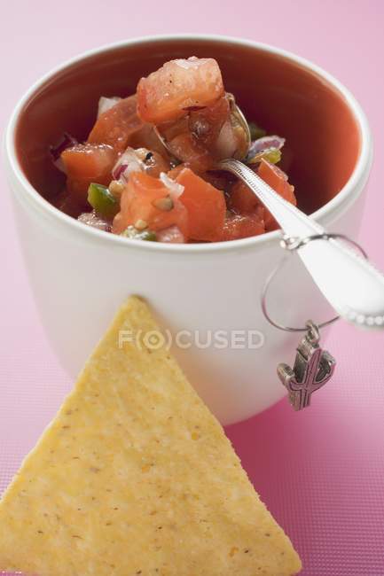 Salsa de tomate em panela com colher, nacho ao lado sobre a superfície rosa — Fotografia de Stock