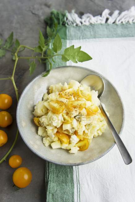 Potato salad with yellow tomatoes on white plate with spoon over towel — Stock Photo