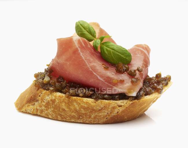 Una rebanada de baguette rematada con hojas de olivo puro, jamón de Parma y albahaca sobre fondo blanco - foto de stock