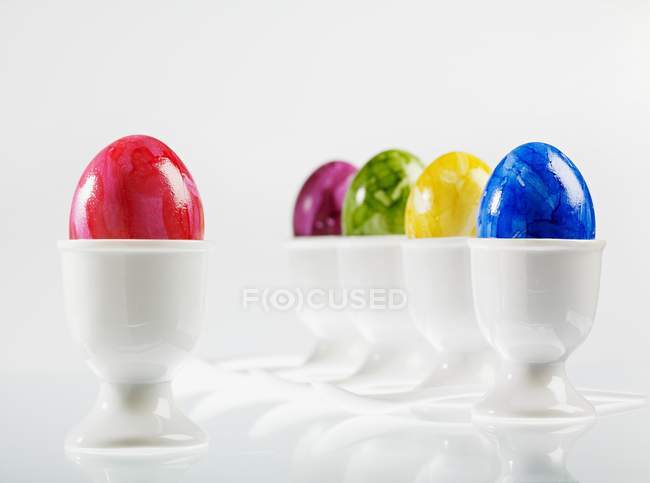 Cinco huevos, de colores brillantes para la Pascua, en copas de huevo blancas con cucharas de huevo - foto de stock