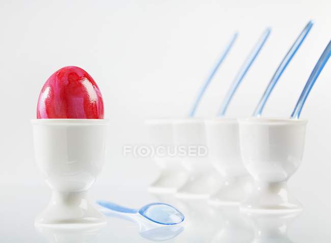 Vista de primer plano de un huevo rojo de color para Pascua junto a una fila de copas de huevo blancas - foto de stock