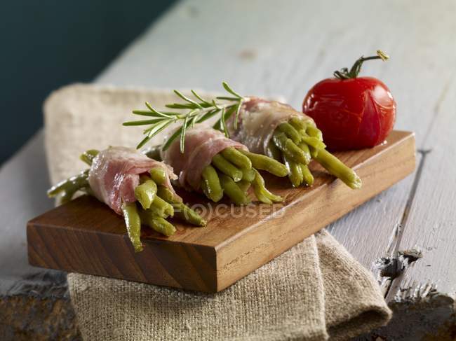 Haricots verts enveloppés dans du bacon avec du romarin sur une petite planche de bois — Photo de stock