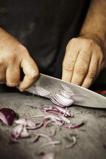 Человек режет красный лук ножом над деревянным столом — стоковое фото
