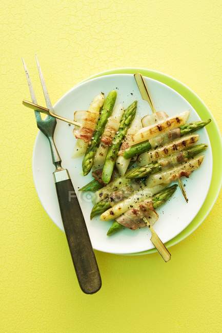Grilled asparagus skewers with bacon on white plate over yellow surface — Stock Photo