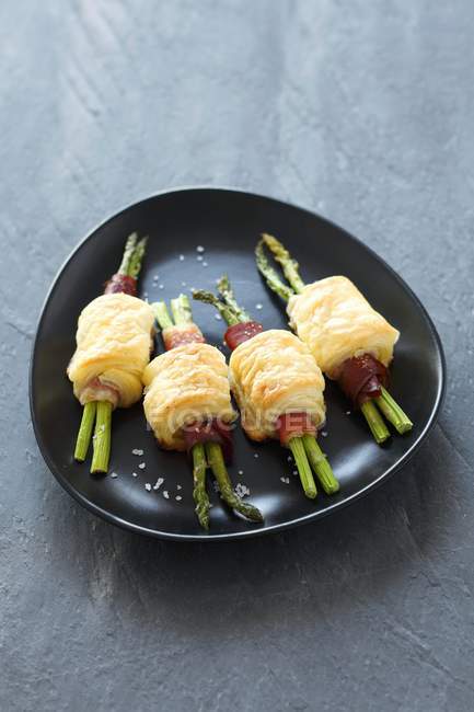Rotoli di pasta sfoglia con asparagi e pancetta su piastra nera su superficie grigia — Foto stock