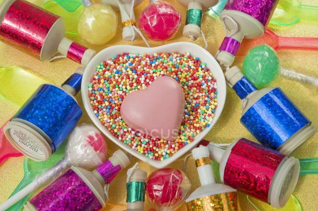 Top view of heart-shaped dish with a pink chocolate heart and colored party poppers and plastic spoons — Stock Photo