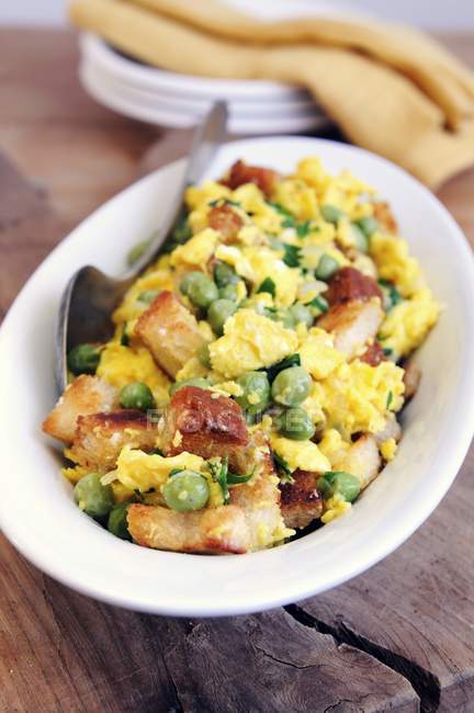 Scrambled egg with fresh peas, Pancetta and croutons on white plate  with spoon on wooden surface — Stock Photo