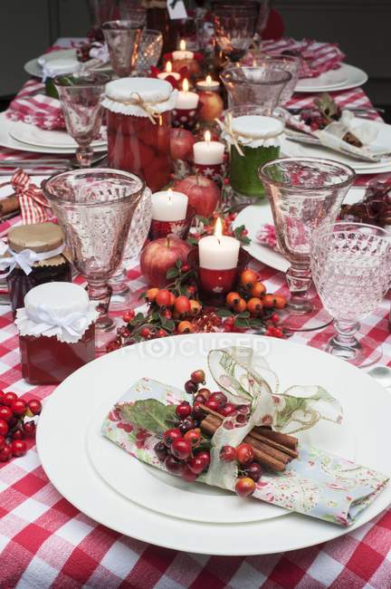 Christmas table laid with a red checkered tablecloth, candles, napkins and jars of jam — Stock Photo