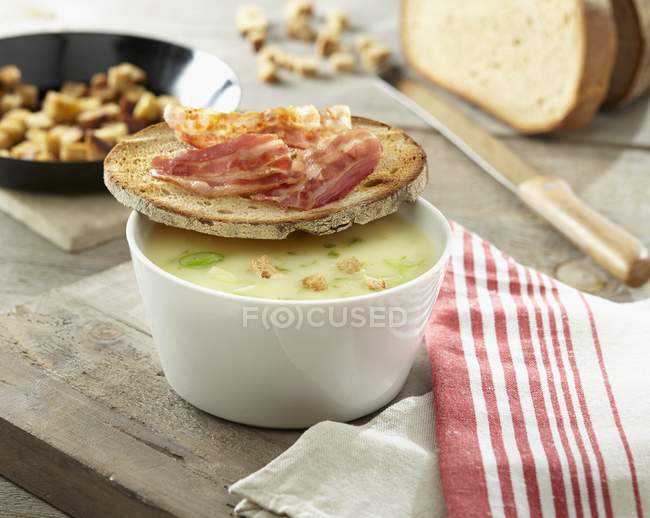 Potato soup with croutons and a slice of bread topped with bacon over wooden surface with knife — Stock Photo