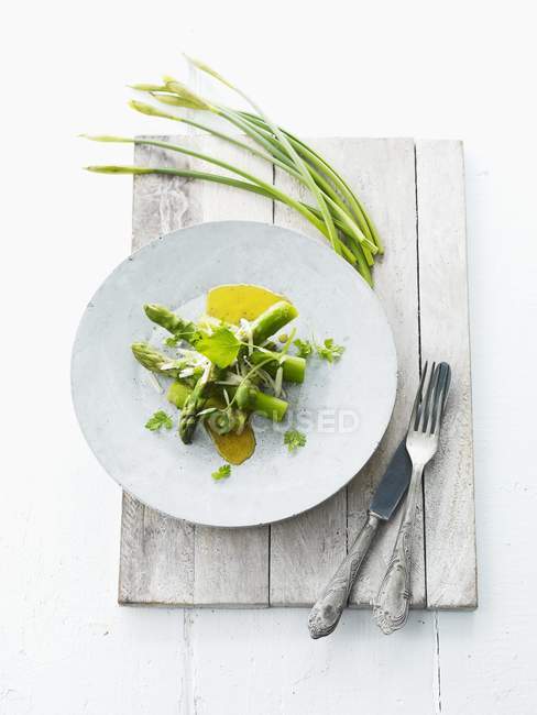 Green asparagus with olive oil and garlic chives for Easter on white plate over wooden surface with fork and knife — Stock Photo