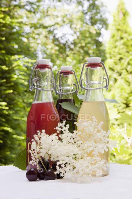 Vue rapprochée de trois bouteilles en verre de sirops maison de rhubarbe, cerise et sureau sur une table de jardin — Photo de stock