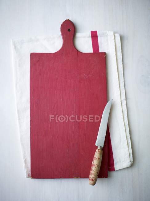 Top view of a red wooden chopping board and a knife on a tea towel — Stock Photo