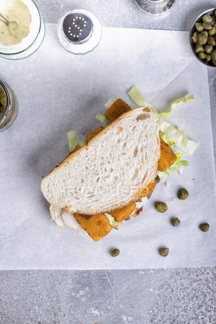 A fishfinger sandwich with tartare sauce and capers laying on paper — Stock Photo