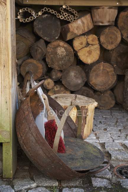 Daytime view of a dustpan and brush in a rusty bowl on cobbles in front of a wood shed — Stock Photo