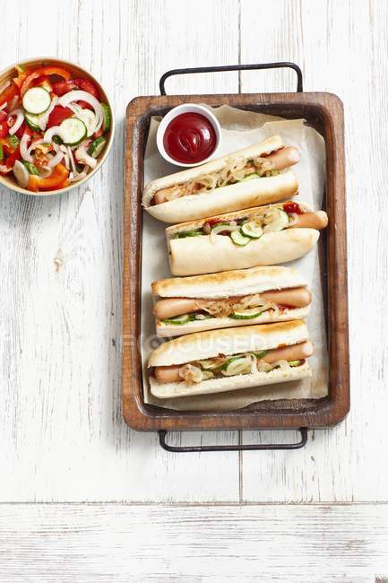 Cachorros-quentes com pepinos, ketchup e cebola apreciam na bandeja sobre a superfície de madeira — Fotografia de Stock