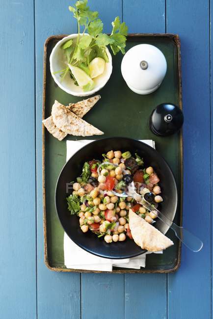 Chickpea salad with tomatoes and olives on black plate over tray on blue wooden surface — Stock Photo