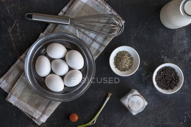 Eier, Gewürze, eine Frühlingszwiebel, Kirschtomaten und ein Schneebesen auf schwarzer Oberfläche — Stockfoto