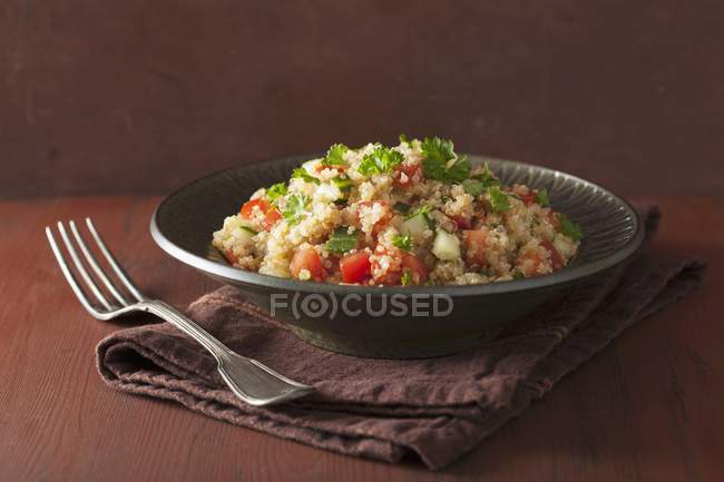Quinoa salad with tomatoes, cucumber and parsley on black plate over towel with fork — Stock Photo