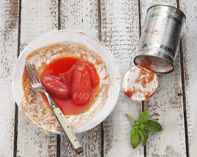 Консервированные помидоры на тарелке с пустой жестянкой рядом с ней на белой тарелке с вилкой — стоковое фото