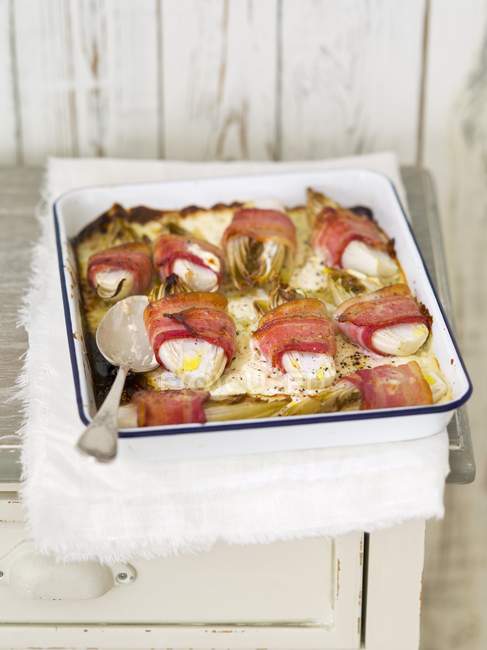 Baked chicory wrapped in bacon in white dish with fork over towel — Stock Photo