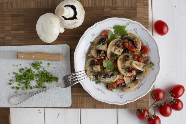 Funghi fritti con pomodori e prezzemolo su piatto bianco su scrivania di legno con forchetta e coltello — Foto stock