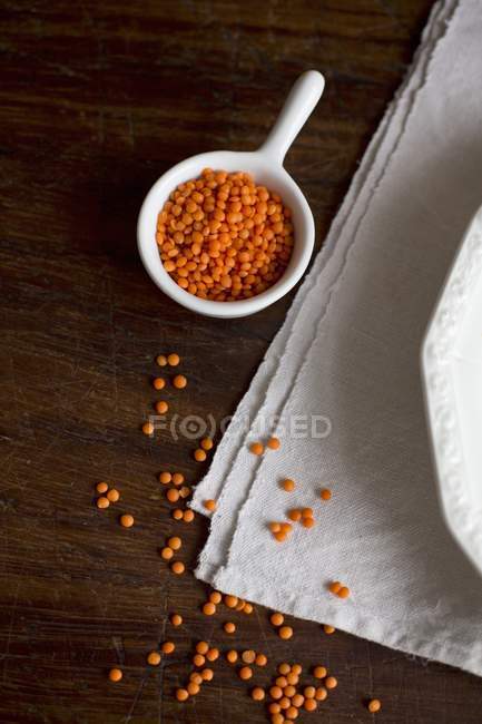 Lentilles rouges dans un bol blanc et dispersées sur le tissu et la surface en bois — Photo de stock