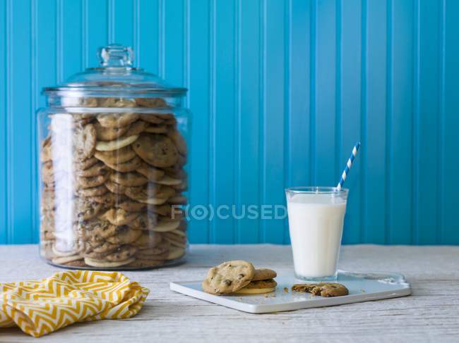Closeup view of cookie jar filled with chocolate chip cookies with plate of cookies and milk — Stock Photo